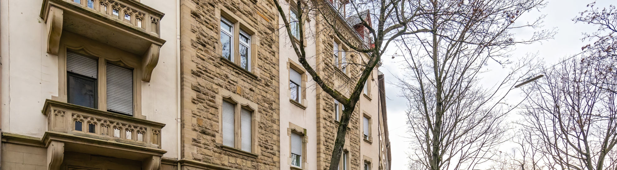 Ein historisches Gebäude mit Steinmauerwerk, Balkonen und Fenstern steht neben kahlen Bäumen unter einem bewölkten Himmel.